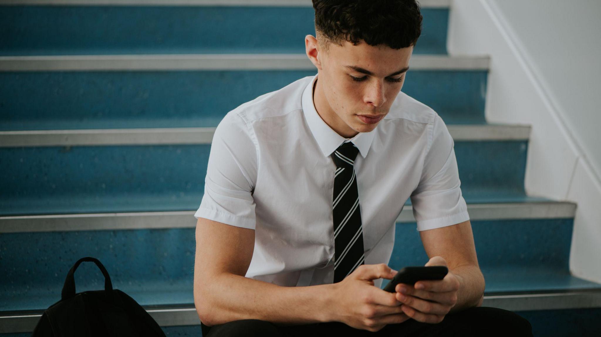 A teenage boy in school, wearing a shirt and tie, sat on some stairs, using his phone