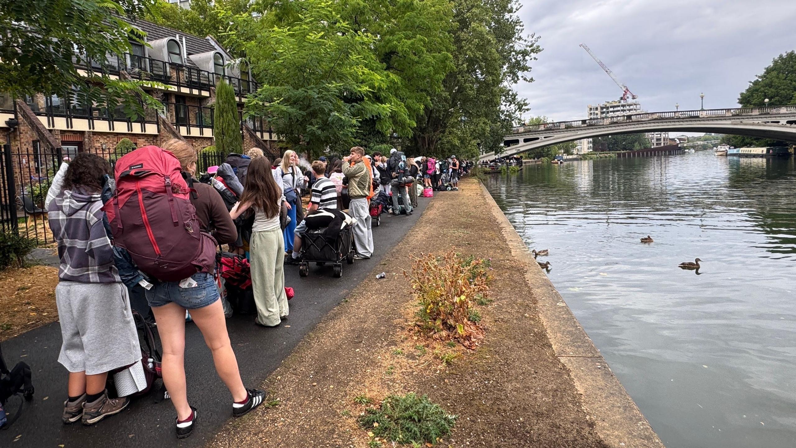 The River Thames is pictured on the right and a long line of people queueing for a boat carrying bags is pictured on the left. The weather is overcast.