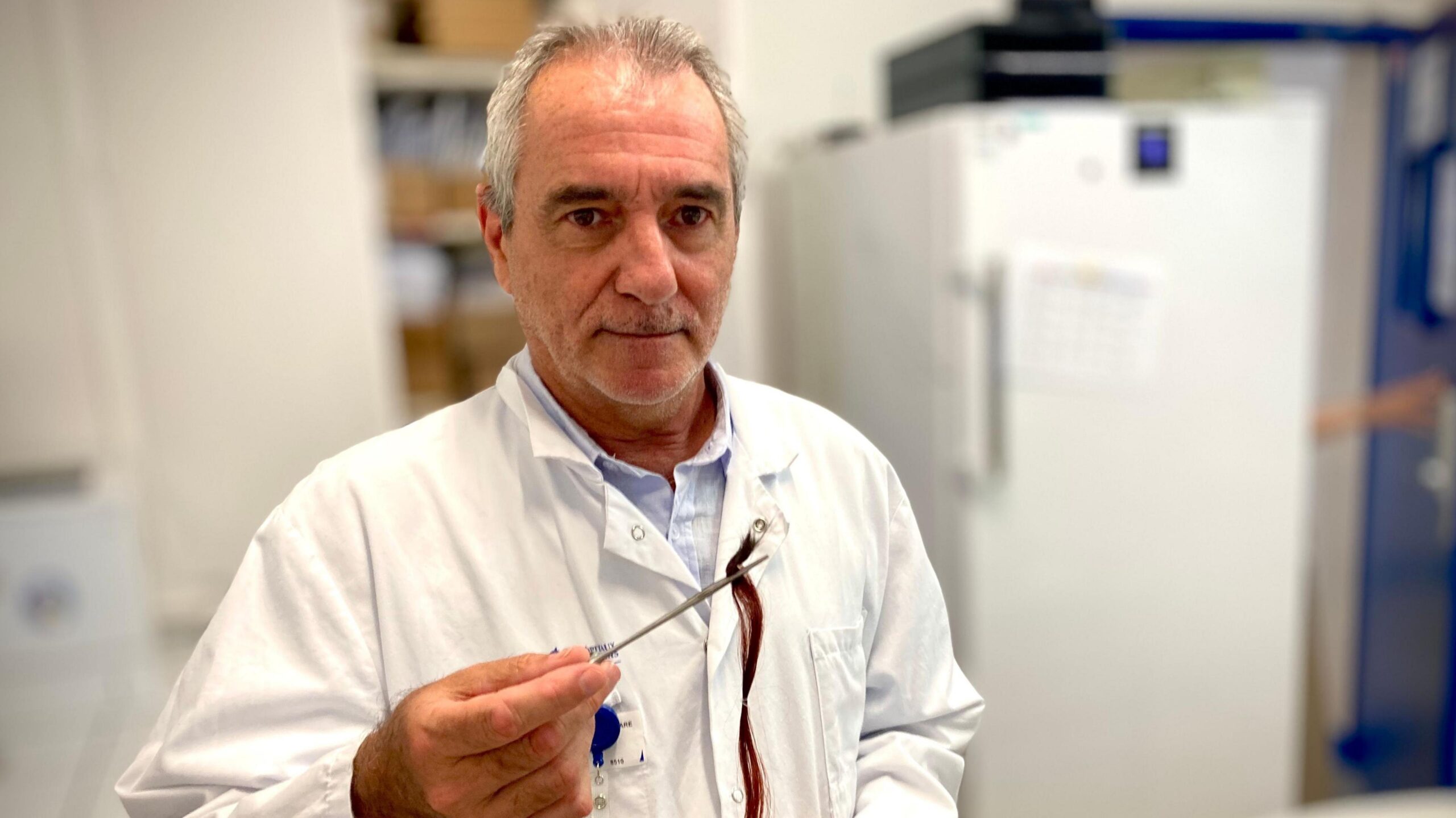Prof Jean-Claude Alvarez is pictured in his toxicology lab wearing a white lab coat. He has short grey hair. He's holding tweezers that are gripping strands of hair