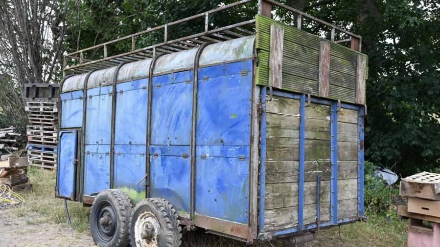A large, enclosed trailer standing on a patch of dry grass and dirt in an outdoor yard surrounded by trees and miscellaneous stored materials.The trailer is painted predominantly blue and has a weathered appearance, with visible rust, dirt and wear on the metal frame. It is mounted on two axles with four large tyres, which are also worn and muddy. The structure is tall and box-shaped, with curved metal roofing panels running along the top. A metal rail framework extends above the roof, creating a raised rack-like structure around the upper edge of the trailer. The side of the trailer is made up of multiple blue metal panels secured by hinges and fasteners. Near the front left side, there is a small access door with a handle. The rear section consists of large wooden panels framed by blue metal supports. The wood is heavily weathered and discoloured, with patches of green staining or moss growth visible across the surface.