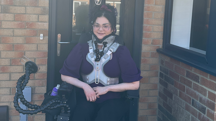 Melanie Hartshorn sits in her motorised scooter outside the front door of her new bungalow. She is wearing glasses, a purple top and black trousers. Her black hair has a purple scrunchie in it and is styled with some hair in a ponytail with some framing her face.