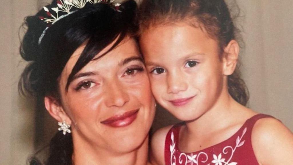 A woman with dark hair tied up with a silver tiara looks at the camera and cuddles a young girl with tied back dark hair and a red dress