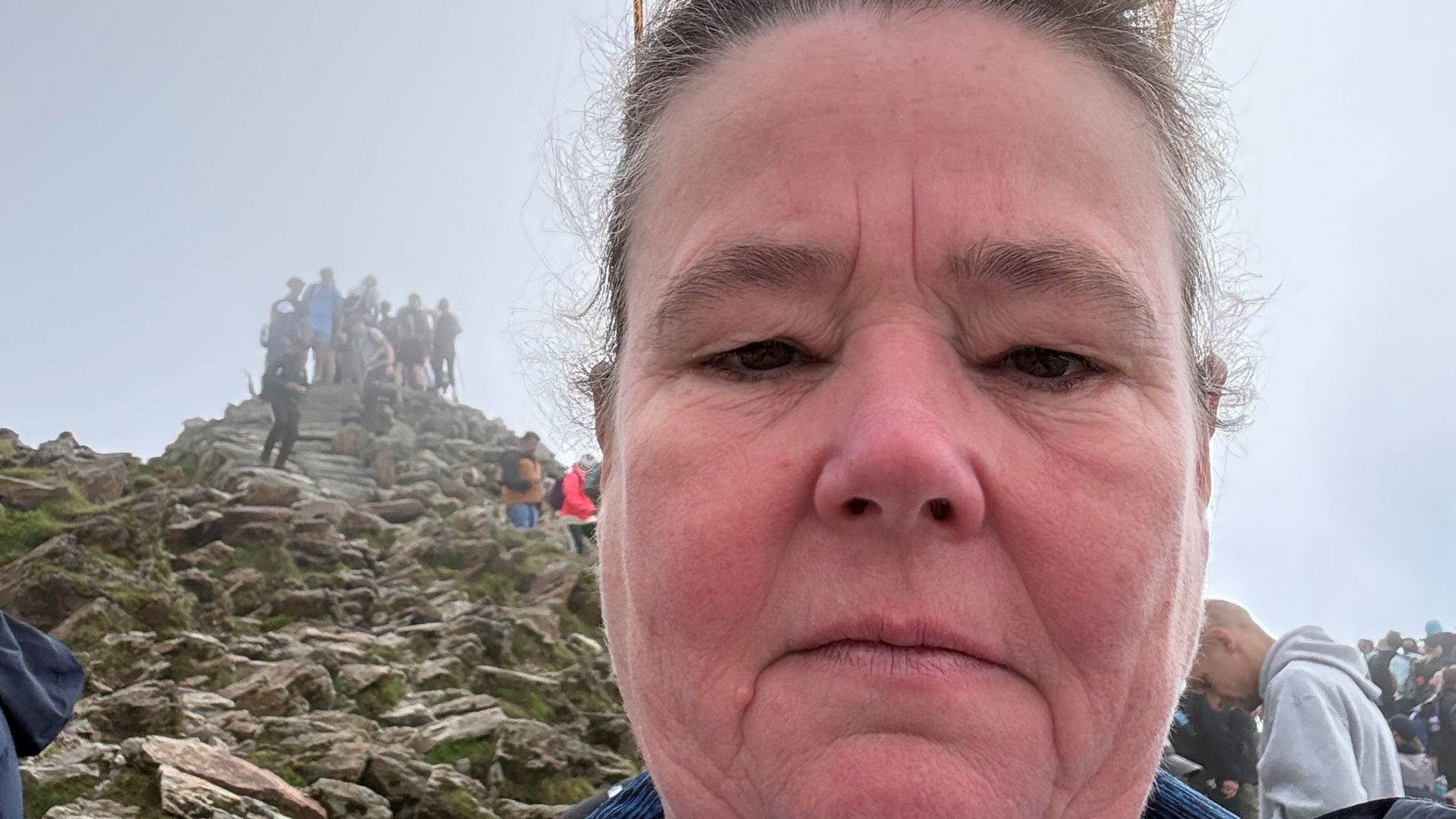 Helen has a neutral expression as she takes a selfie overlooking the summit of Yr Wyddfa