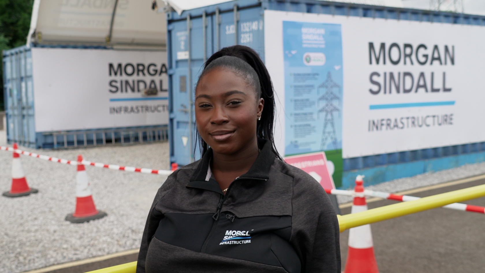 A young woman  in a black Morgan Sindall Infrastructure zip-up jacket standing in the foreground at a construction or infrastructure site. Behind the person are two blue site cabins or container units displaying large Morgan Sindall Infrastructure signs. Red-and-white traffic cones and barrier tape mark off a gravel area, with a yellow safety barrier running across the lower right of the image. Additional site signage is attached to one container, and electricity pylons are visible in the background.