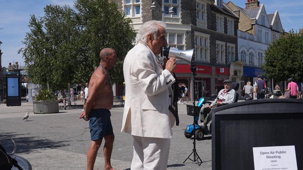 Erlam wears a white suit and is speaking into a mega phone