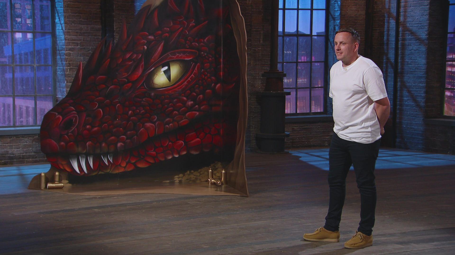 Nathan Murdoch is standing in a studio wearing a white T-shirt and dark trousers. There is a large painting of a scaly, red dragon with yellow eyes next to him.
