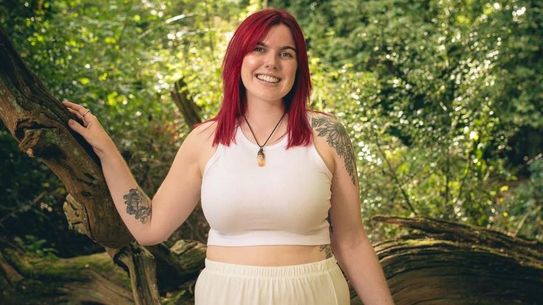 A young woman wearing a cream crop top and skirt. She has bright red hair and tattoos. She is stood in front of a tree.