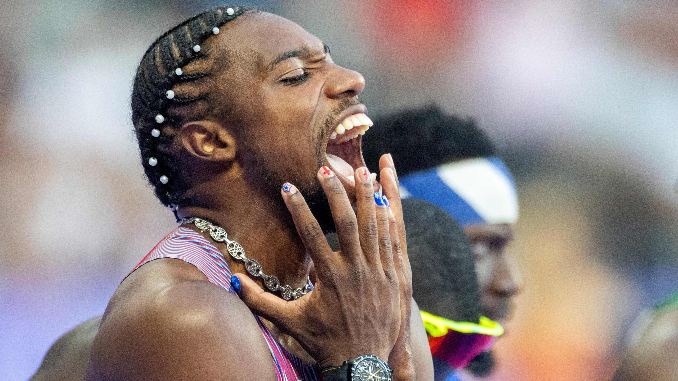 US athlete Noah Lyles' mouth is open in a yell as he holds up his hands to his face, and his nails are painted in red and blue designs.