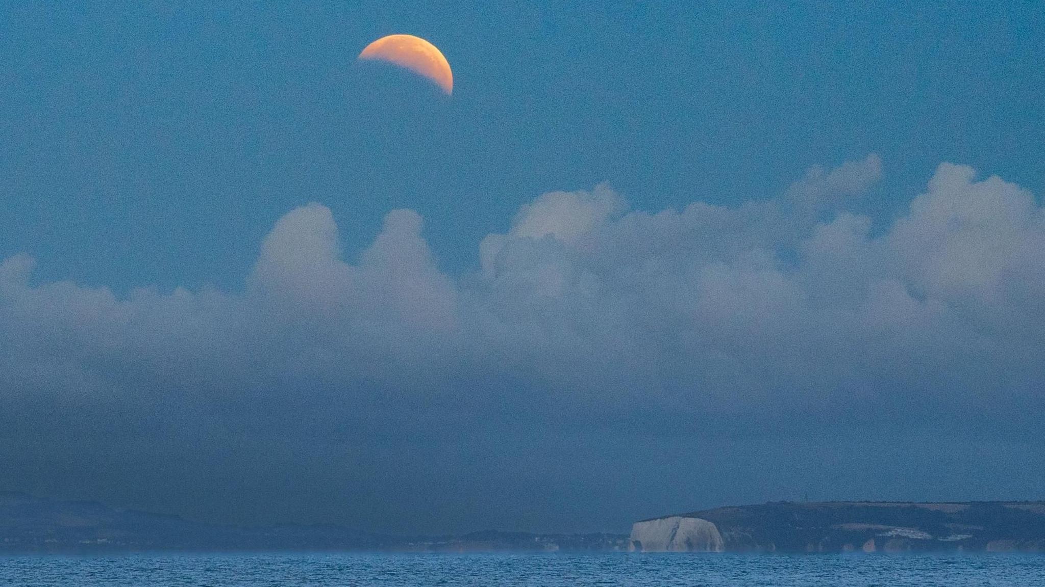 Coastal view showing distance white cliffs, some billowy cloud above, with the crescent of a partially eclipsed moon
