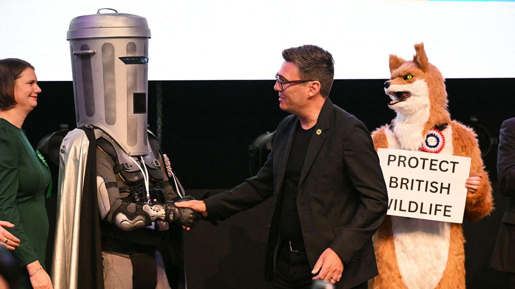 Burnham is in the centre of the photograph, wearing a black suit over a black t-shirt, and shaking hands with Count Binface on his left. To his right is a man dressed as a fox, holding a placard reading Protect British Wildlife.
