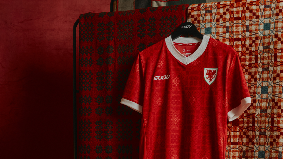 A red Wales football shirt hangs on a black hanger in front of two patterned blankets. The jersey features white trim on the collar and sleeves, the Wales crest with a red dragon on the left chest, and the SUDU logo on the right. The fabric includes a subtle tonal pattern, with warm red lighting throughout the image.