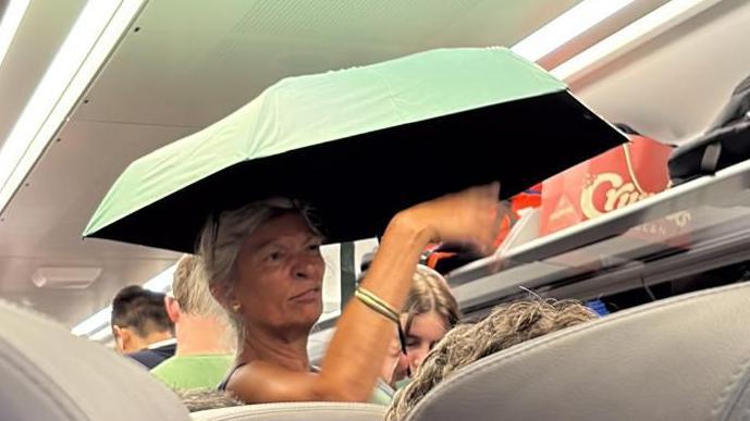 A woman holding an umbrella whilst standing in the aisle of a busy train. Other people are standing and the overhead shelf is packed with bags.