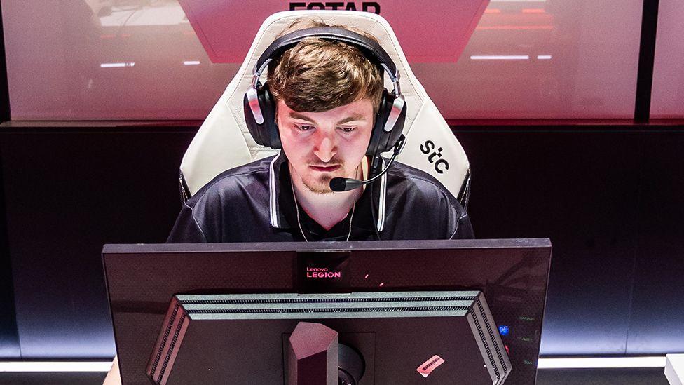 A young man wearing a look of intense concentrations sits in front of a black computer monitor. He's wearing a large, expensive-looking gaming headset with a microphone attachment, and sitting in a large, white gaming chair.