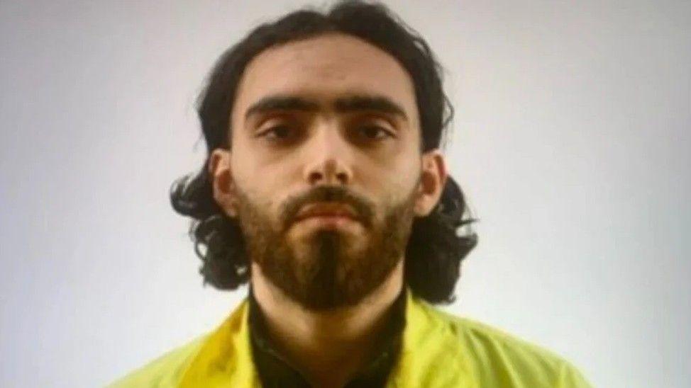 A man with long dark hair and a short beard in a mugshot-style photo