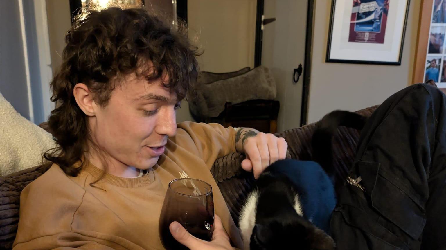Luke Grindlay, a young man, with long, dark curly hair, is sitting on a sofa looking down and smiling at a black and white cat sitting on his lap.