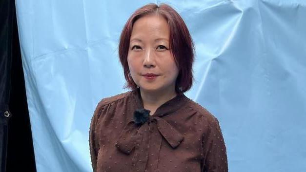 Sachie Mikawa stands in front of a blue tent wearing a brown top with a bow tied at the neck