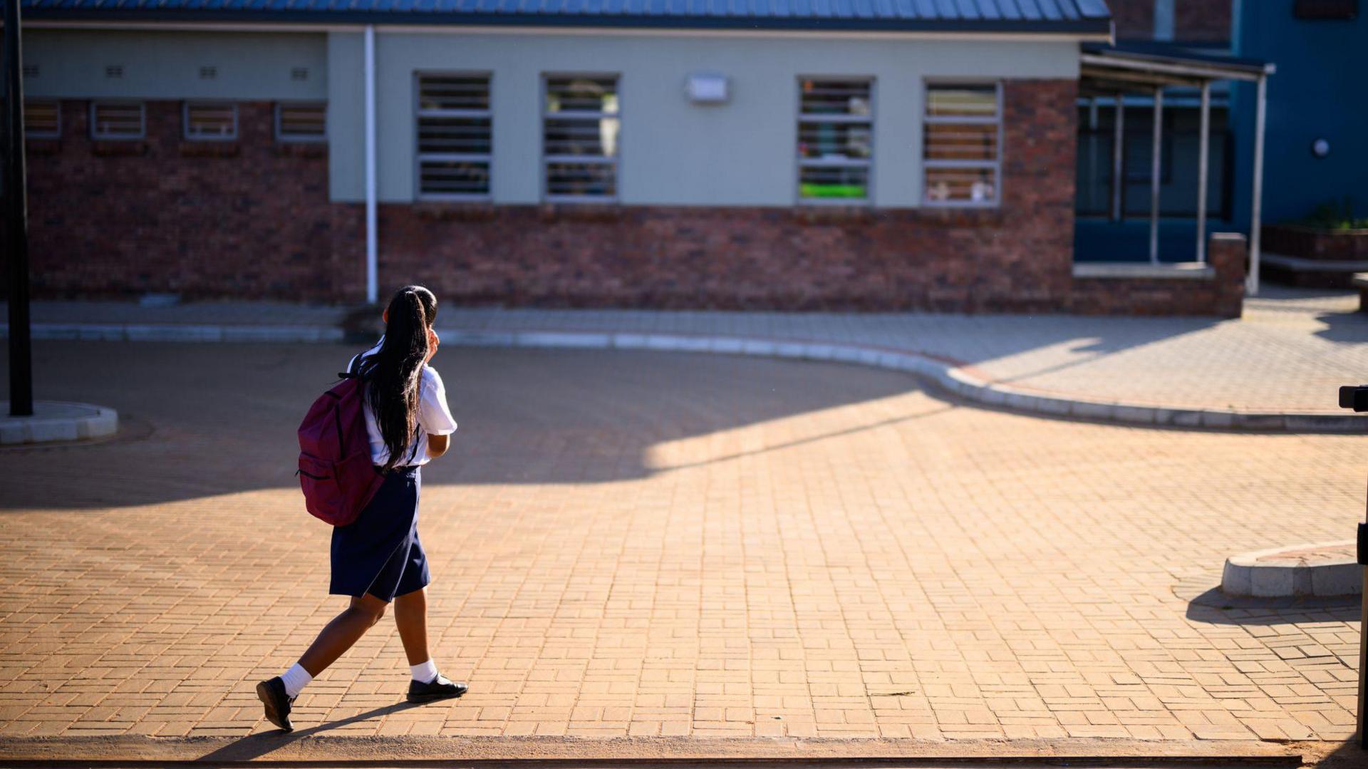 A child carrying a large red backpack walks across a paved school courtyard towards low-rise school buildings. The scene is brightly lit, with long shadows and brick-and-render buildings visible in the background.