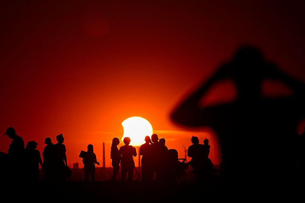 An orange sky with the Sun in the centre, there is a small bite sized chunk taken out of its left side as the Moon covers the surface. People standing around in groups with telescopes are in shadow