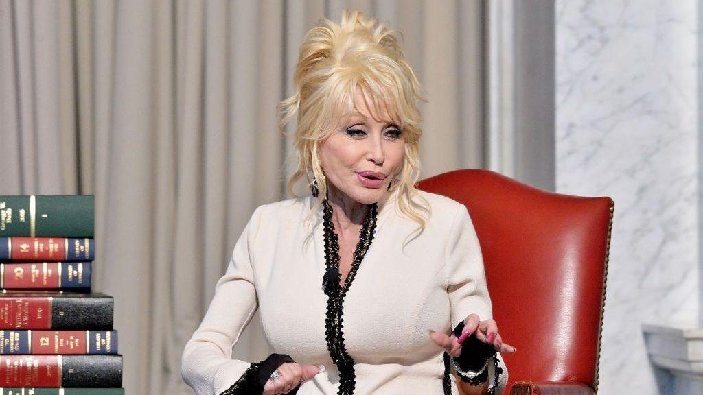 Dolly Parton, wearing a black and white blazer, sits on a red leather chair and speaks to a crowd. There is a pile of old-style books next to her.