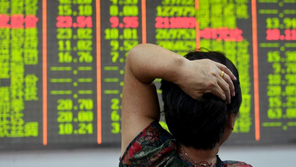 An investor looks at an electronic board showing stock information with numbers highlighted in red and lime green against a black background. The investor has her back to the camera and has her left hand resting on her head.