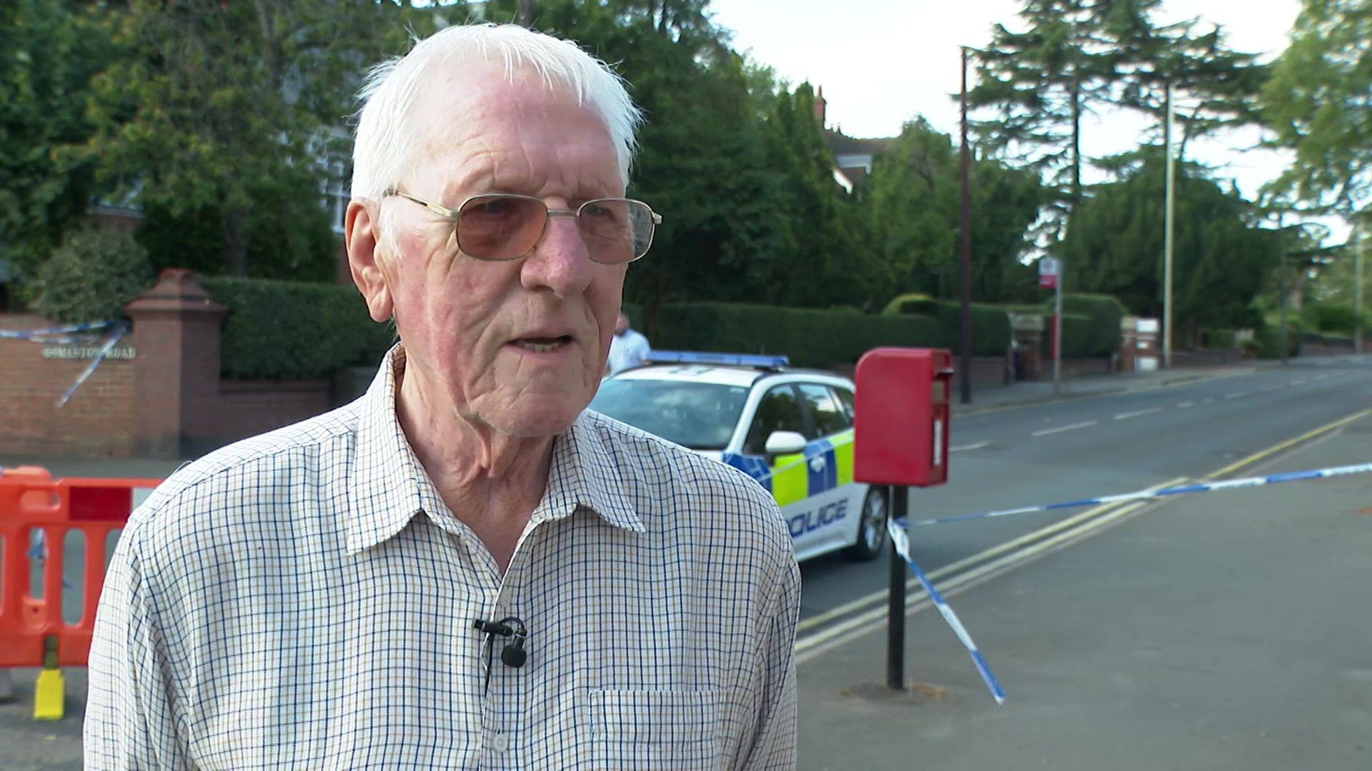 A man stands in the street with cordon and police tape behind him. He has white hair and wears glasses.