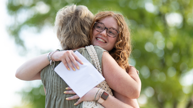 A girl with curly red hair and glasses hugs a woman with short blonde hair. The girl is smiling and holding a white piece of papers. You can't see the other woman's face but she wears a green top and beige bag. Behind them are green trees.