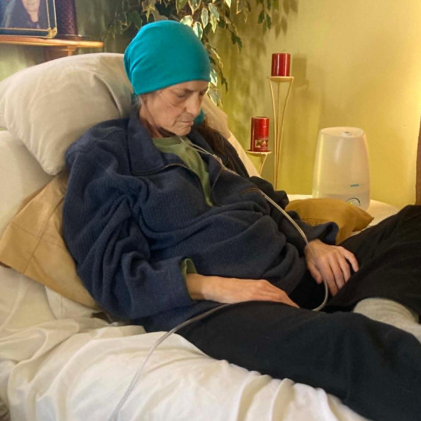 Kelly lies on a hospital bed propped up by pillows and looking down, as if asleep. She wears a blue headscarf on and has a clear tube feeding into her nose. Her hands rest of her lap.