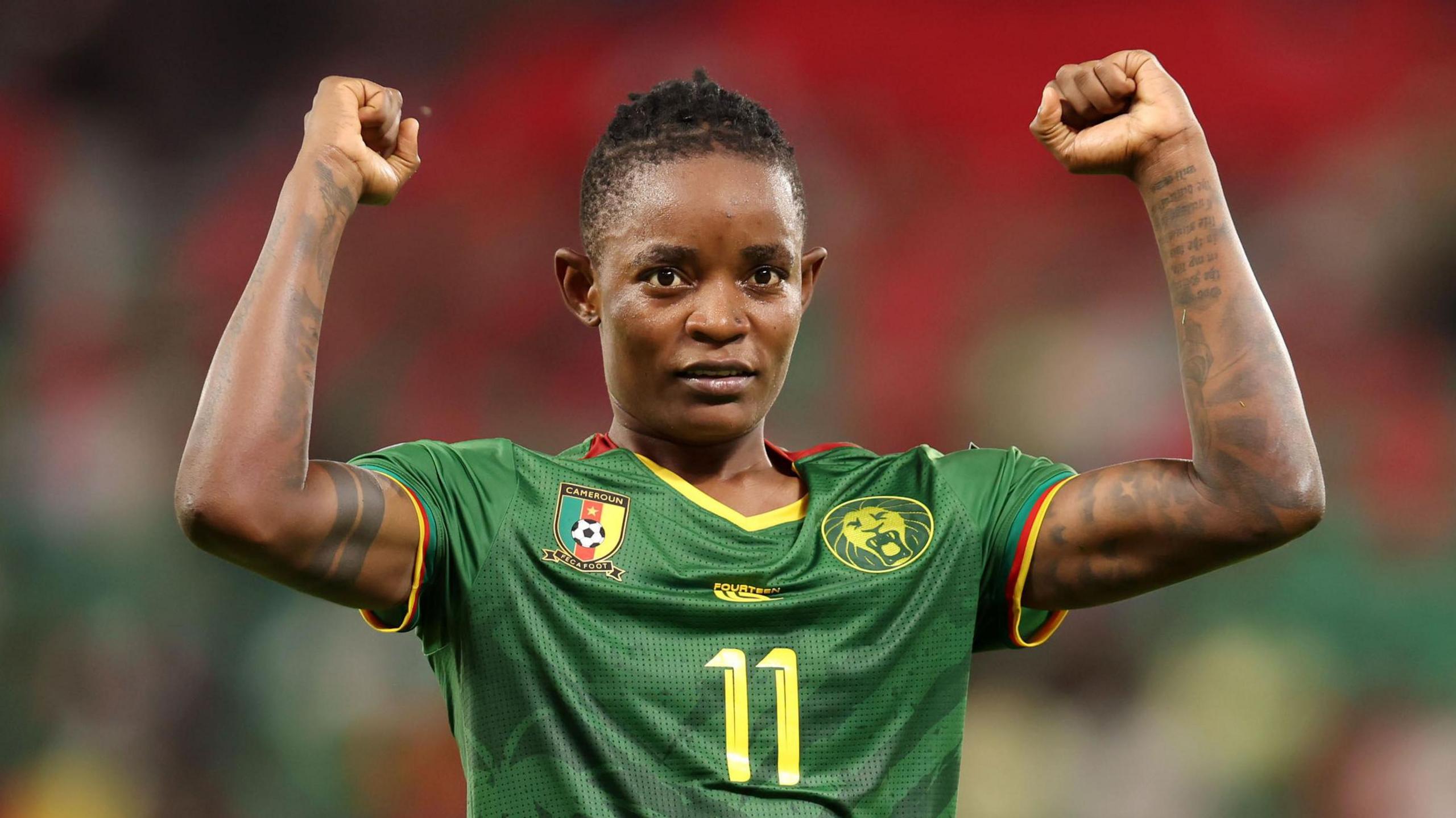Marie Ngah Manga, wearing a green Cameroon shirt with a yellow number 11 on the chest, holds up both arms with fists clenched in celebration