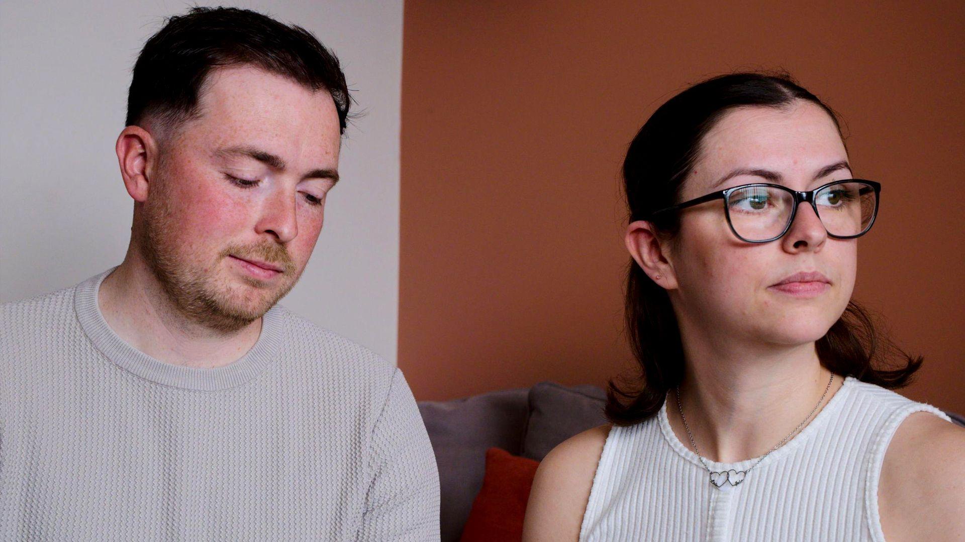 The McCreadys are sitting together on a sofa. Lauryn is looking wistfully out of a window while Andrew is looking downards towards their daughter's memory box