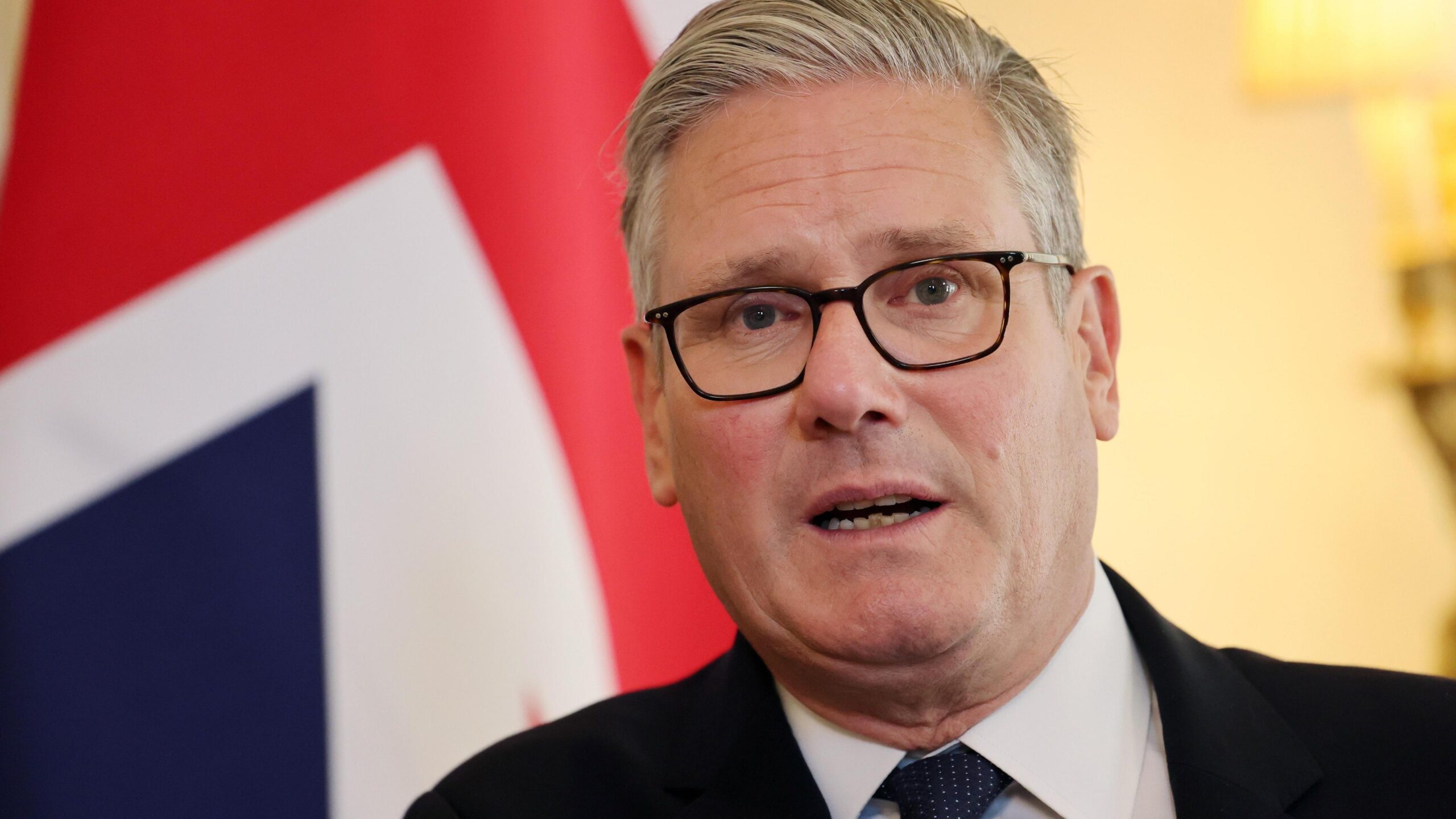 Sir Keir Starmer speaking in front of a union jack flag. It is a head and shoulders shot - he is wearing a white shirt, dark black suit and a dark tie.