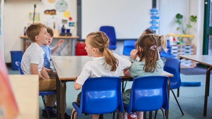 Stock image of young school pupils. They are sitting at a desk and wearing summer uniform. They have blue chairs and behind them is a smartboard, teachers' desk and green plant.
