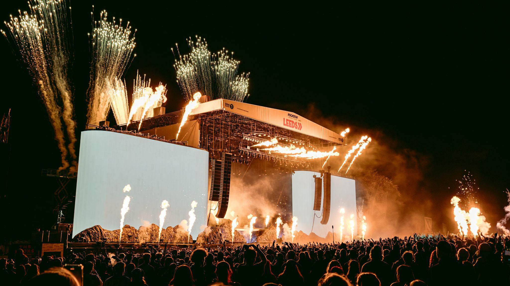 Fireworks light up the sky behind a large stage. A huge crowd of people can be seen lit up in the light from the fireworks.