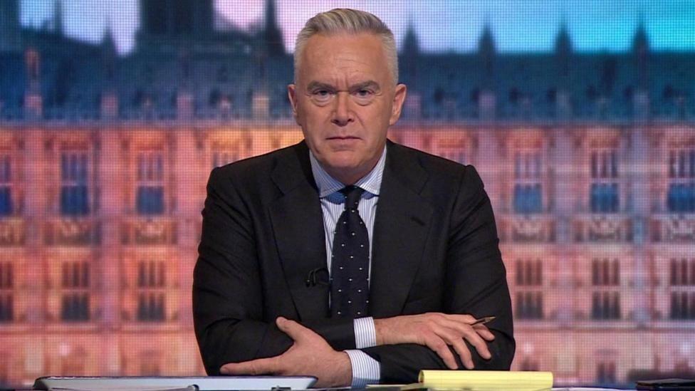 Huw Edwards, who has grey hair, wearing a dark jacket and blue tie over a light shirt