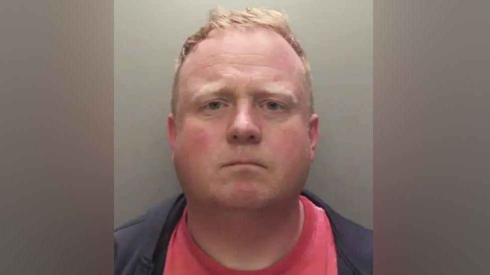 Robert Bush with short, light brown hair is shown in a straight‑on mugshot photograph. He has a neutral expression and is wearing a dark jacket over a red shirt. The background is plain and grey.