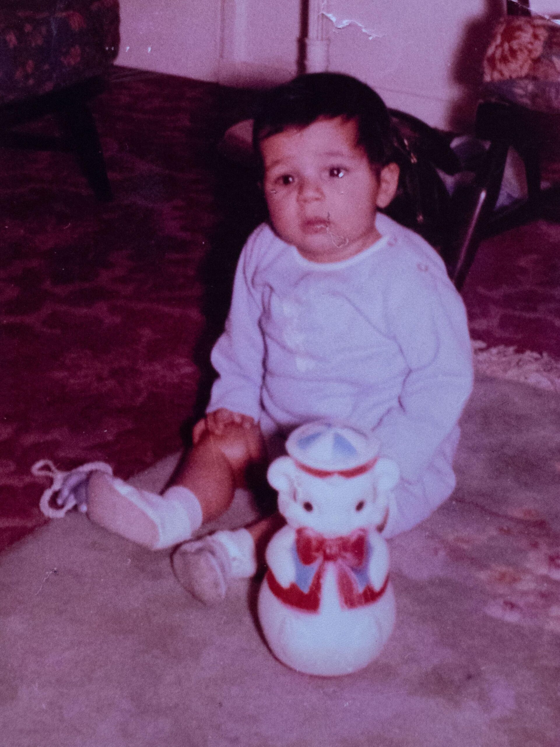 An old photograph of a baby boy sitting on the ground with a toy. He is in a baby-blue outfit and has a full head of dark hair.