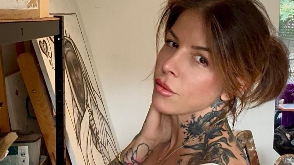 A woman is sitting down and looking at the camera posing, showing her tattoos on her neck and wrist.