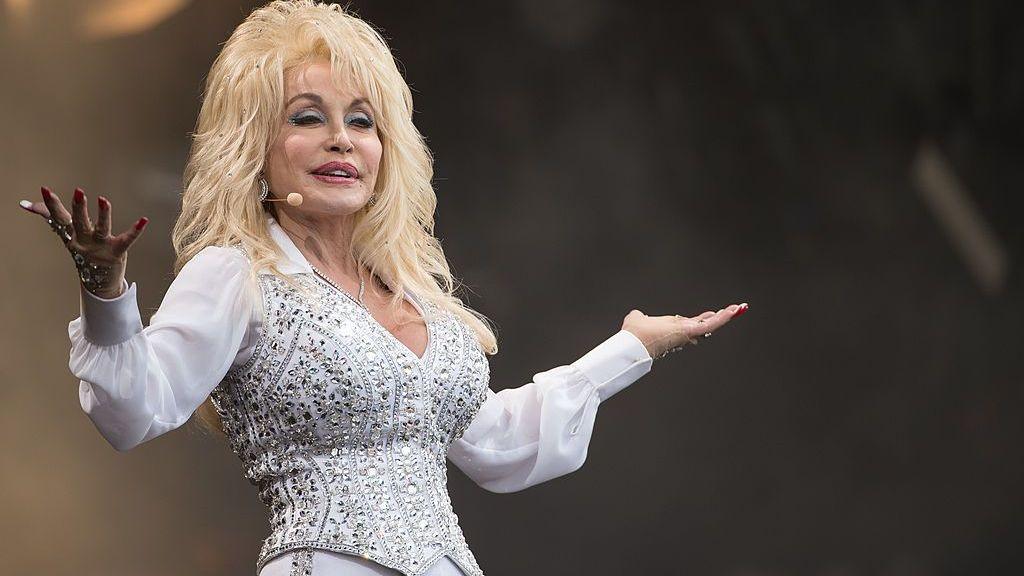 Dolly Parton, wearing a white and silver bedazzled outfit, on stage smiling and holding out her arms