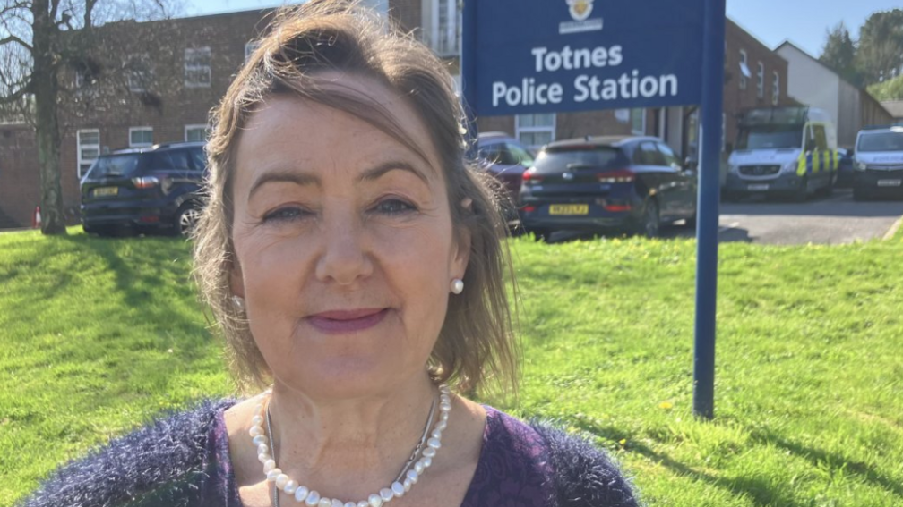Joanna Brittan looking at the camera and smiling. She has a short dark bob and is wearing a pearl necklace and earrings. She also has on a purple top. She is pictured from the shoulders up. There is a grass back behind her with a sign on it saying "Police Station".