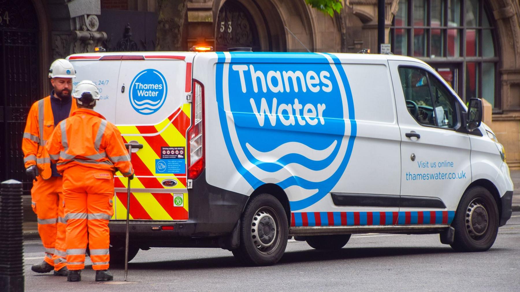 A thames water van parked on a streert with two workers dressed in orange overalls standing to to the left of the van