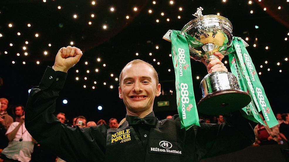 Graeme Dott after winning the World Snooker Championship in 2006. He is holding his trophy in the air. It is silver and has green ribbons attached.