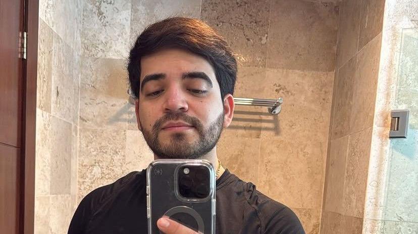 César Gastélum looks into a mirror as he takes a selfie. He has a beard and black hair. He is wearing a black T-shirt and a gold chain around his neck