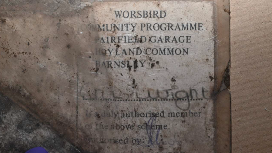 A worn and heavily stained paper card or document inside what appears to be a transparent plastic sleeve. The document is lying on a rough wooden or cardboard surface and is covered with dirt, scratches, dark marks, and signs of age, making much of the text difficult to read. The card contains printed text in black capital letters. The upper section appears to read: WORSBIRD  COMMUNITY PROGRAMME  AIRFIELD GARAGE  HOYLAND COMMON  BARNSLEY Some letters are obscured by dirt and damage, so parts of the text are difficult to confirm with complete certainty.