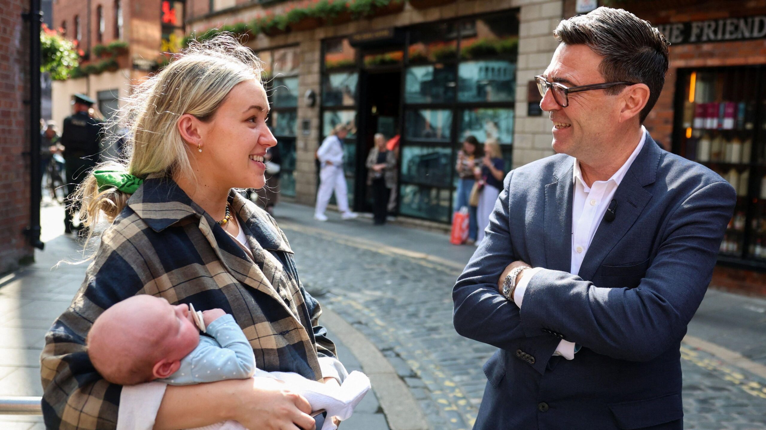 Prime Minister Andy Burnham speaks with a woman in Belfast, as he makes his first visit to Northern Ireland since coming to office. He has dark hair, wearing glasses, a navy blazer and white shirt. His arms are folded. The woman has long blonde hair tied back, wearing a check coat and carrying a baby.