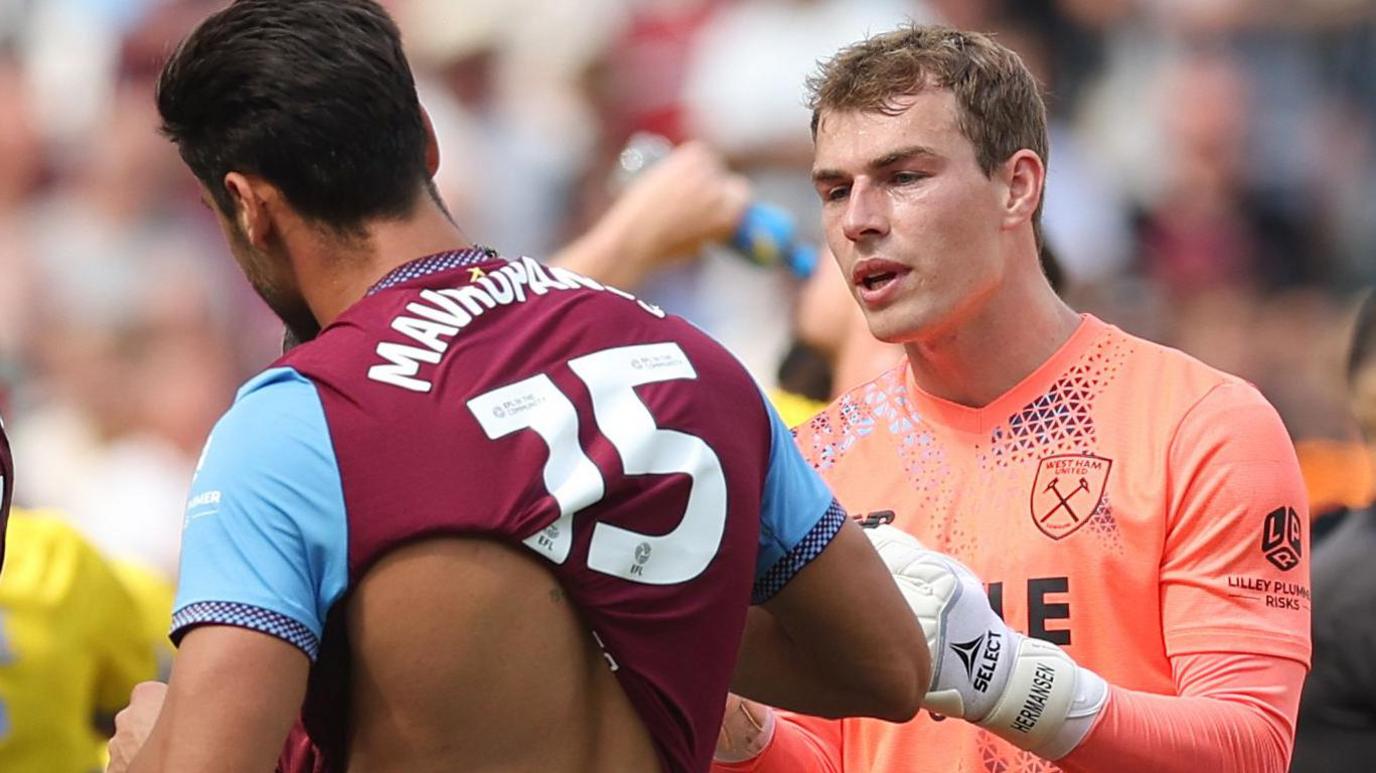 Mads Hermansen, wearing West Ham's orange goalkeeper shirt, speaks to his team-mate Konstantinos Mavropanos, who is wearing the claret and blue home shirt and has his back to the camera
