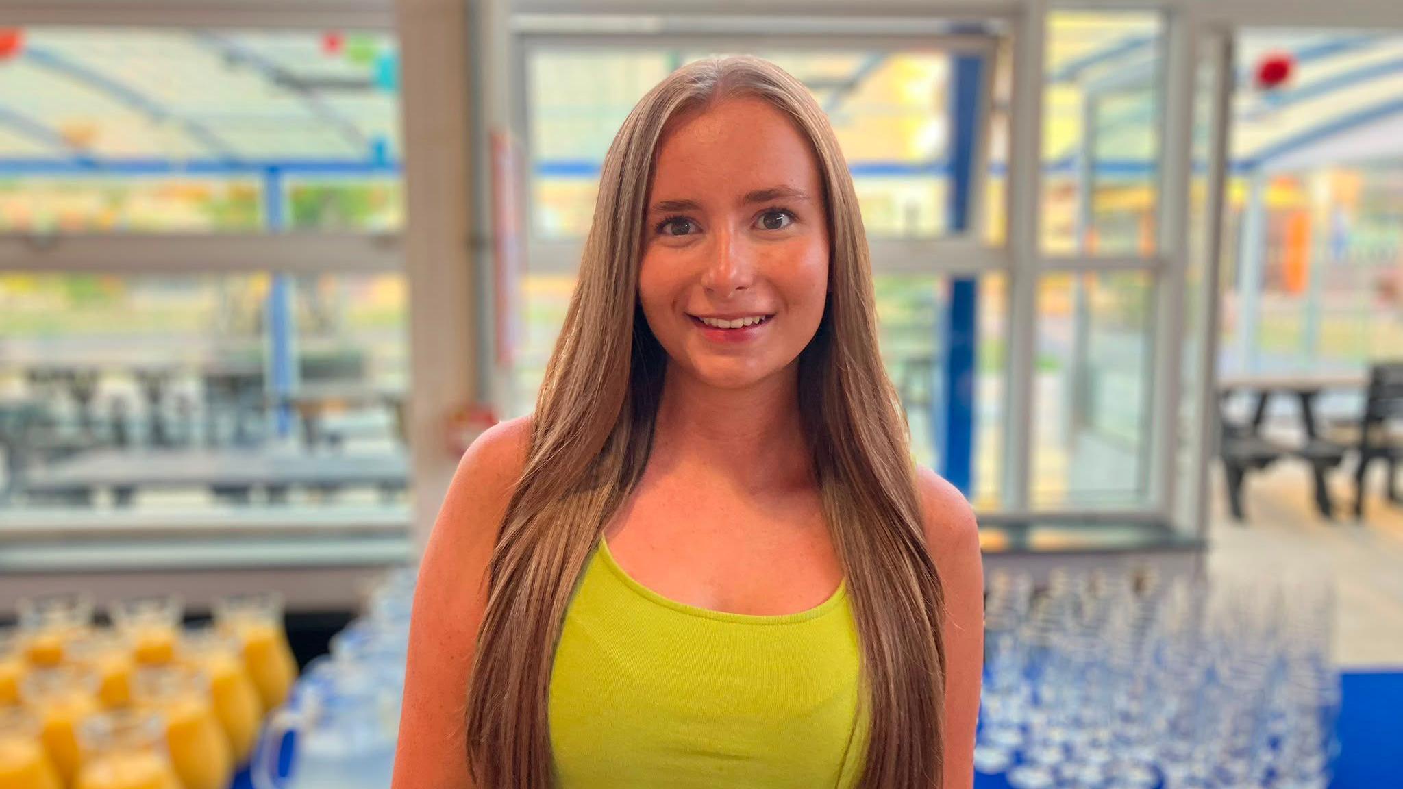 Niamh wears a yellow top and is smiling. She is standing in front of a table with glasses and orange juice on