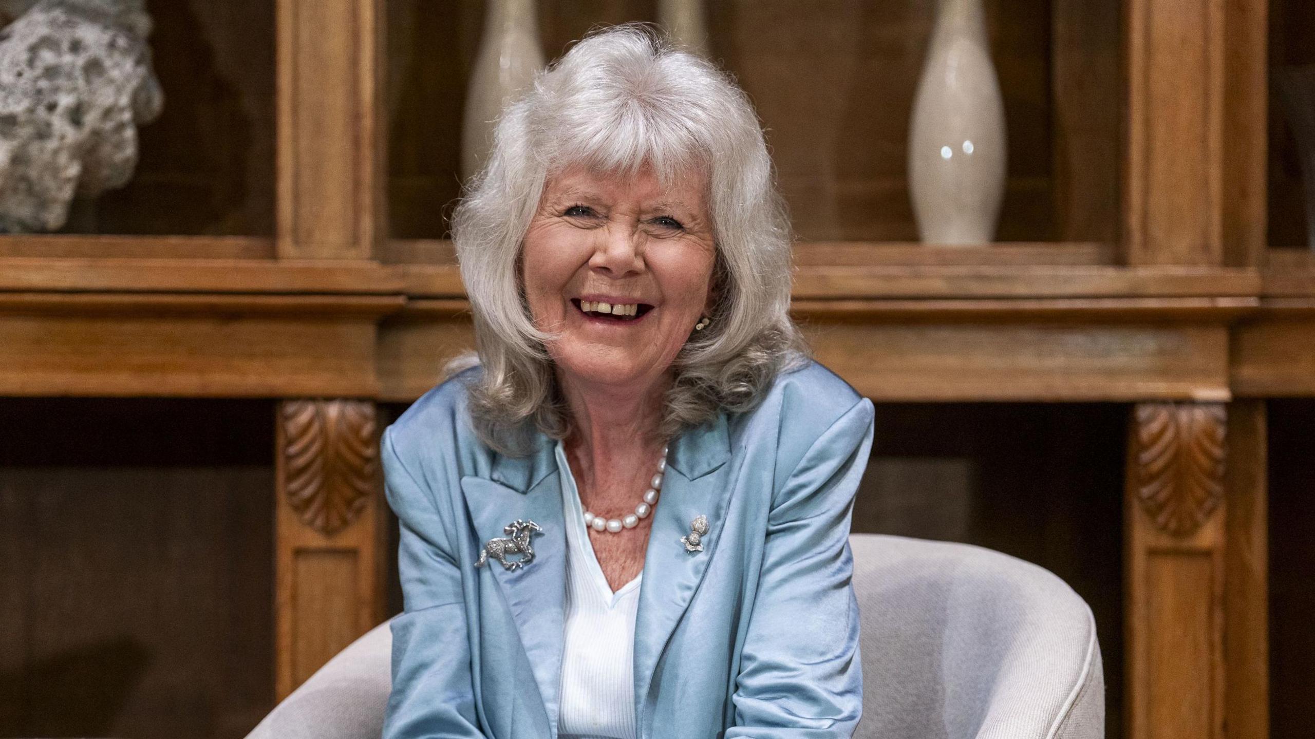 Dame Jilly Cooper, who is wearing a satin blue blazer with a horse brooch on her lapel, a pearl necklace, and a white v-neck top, laughs as she sits on a grey chair. She has shoulder-length grey hair and a fringe. She is sitting in front of a wooden panelled wall with vases and ornaments on the shelf.
