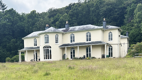 Dol-llys Hall taken from the far side of a long-grass lawn. The hall is painted light yellowy cream and has a slate tiled roof. To the left and right of a round tower-like protuberance are two covered verandas with supporting posts. Six large arched white-framed leaded windows are visible on both floors, with three chimney sets on the roof. The house is framed by large, mature woods. Some washing is visible on a line to the right of the picture.