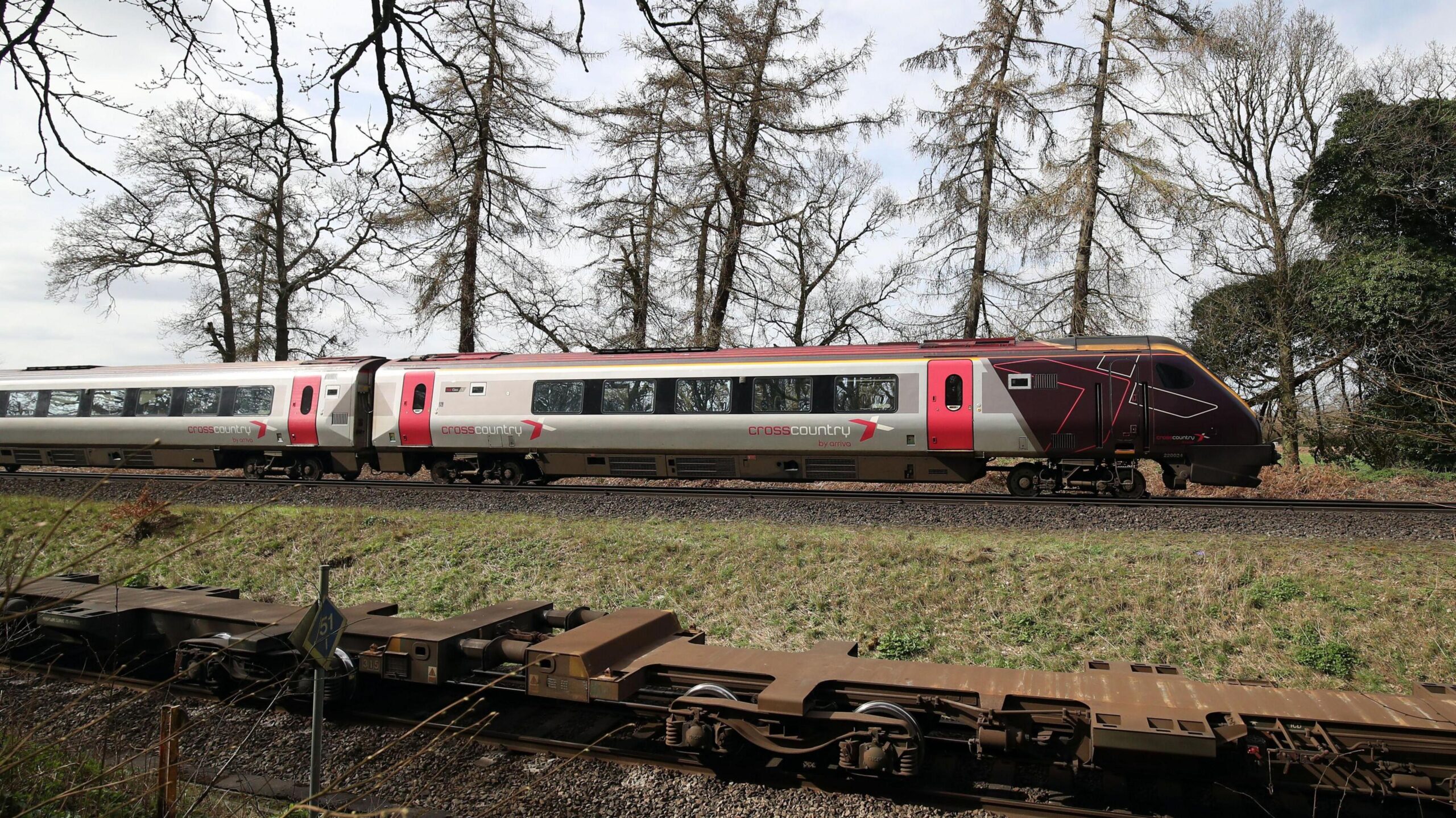 The cab and first carriage of a CrossCountry train travels on tracks in the countryside. In the background trees and fields can be seen.