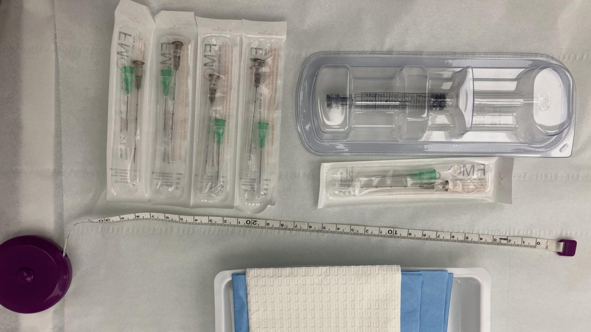 The image shows a neatly arranged set of medical supplies laid out on a sterile-looking white surface. Several individually packaged cannulas are positioned on the left, while a larger pre-packaged syringe sits in a clear plastic tray on the right. A measuring tape stretches horizontally across the middle of the image, and a tray containing absorbent pads or dressings is visible near the bottom edge.