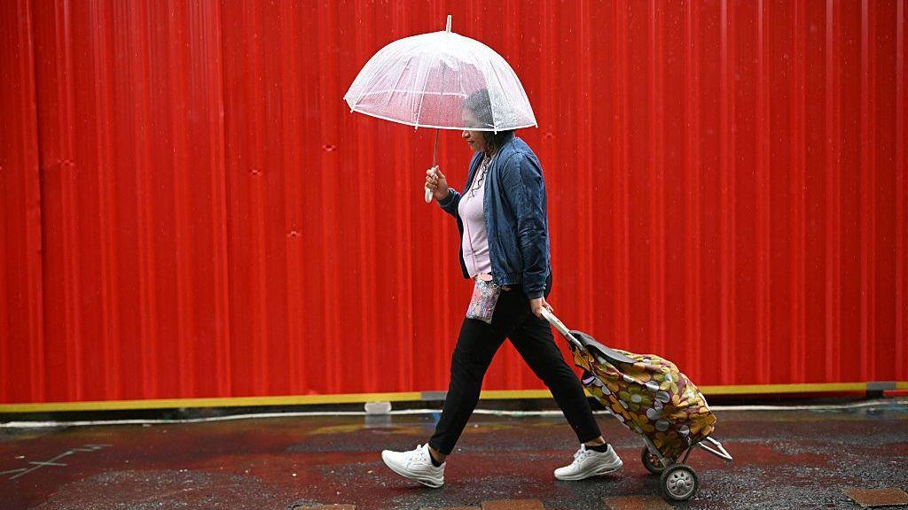 A woman walks along in the rain pulling a shopping trolley